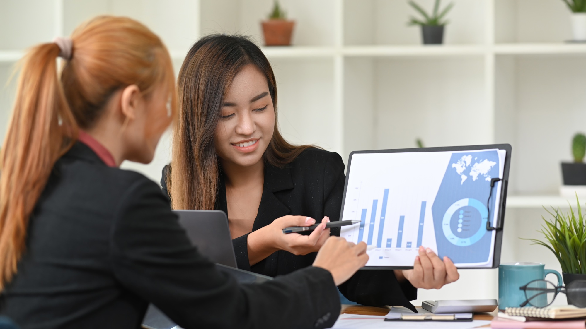 Two business women analyzing marketing research reports together at office desk.
